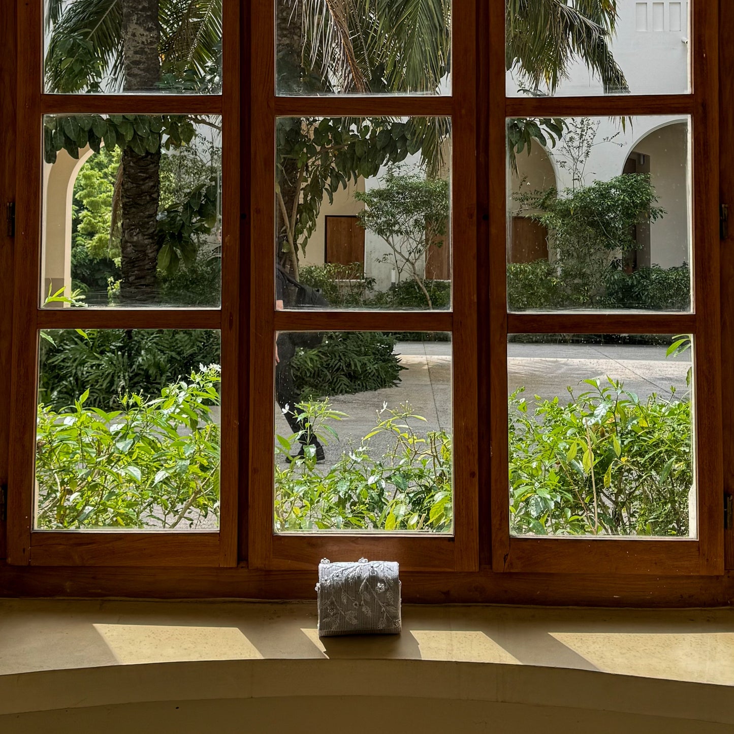 White aso-oke bag framed in an arched window with wooden frame showing a view of palm trees and greenery outside.